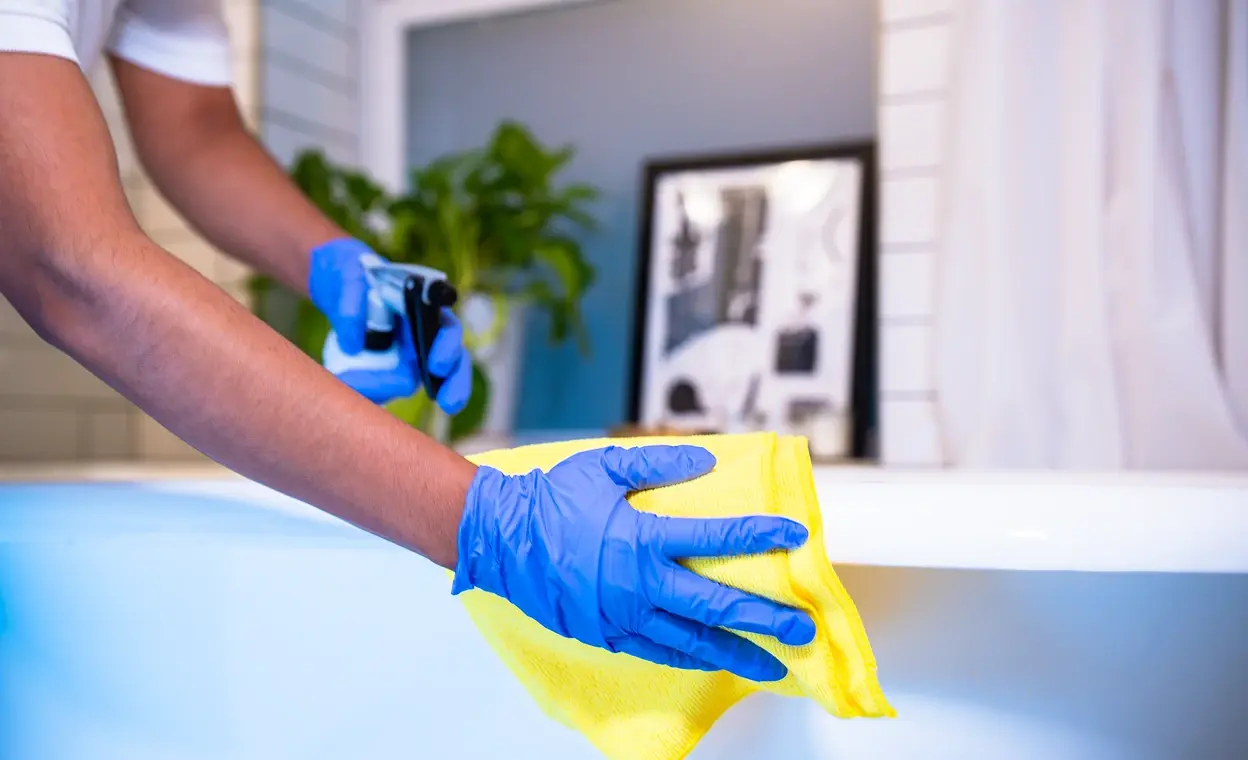 Professional cleaner wearing blue gloves, wiping a bathtub with a yellow cloth in a spotless bathroom, symbolizing care and attention to detail