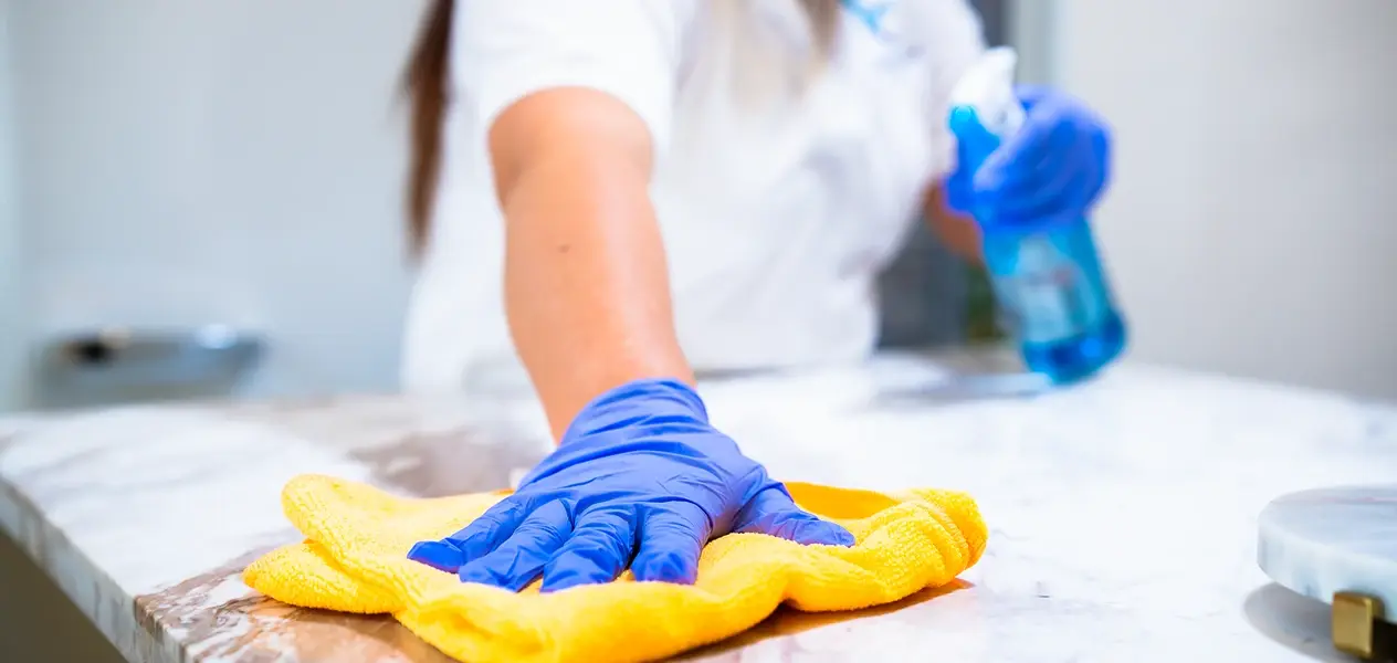 Professional cleaner wearing blue gloves wiping a marble countertop with a yellow cloth and spray bottle in hand