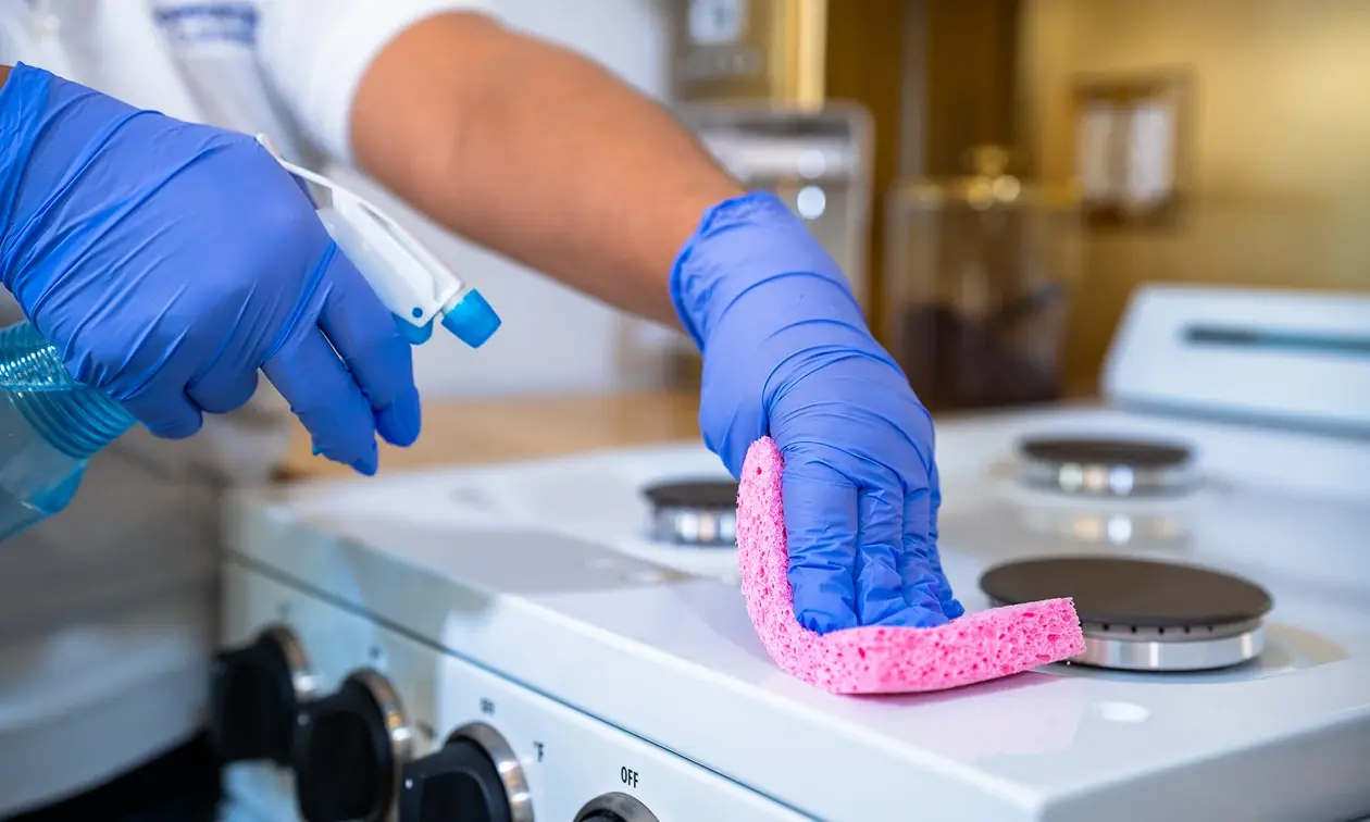 Professional cleaner wearing blue gloves, scrubbing a stovetop with a pink sponge and cleaning spray in a modern kitchen