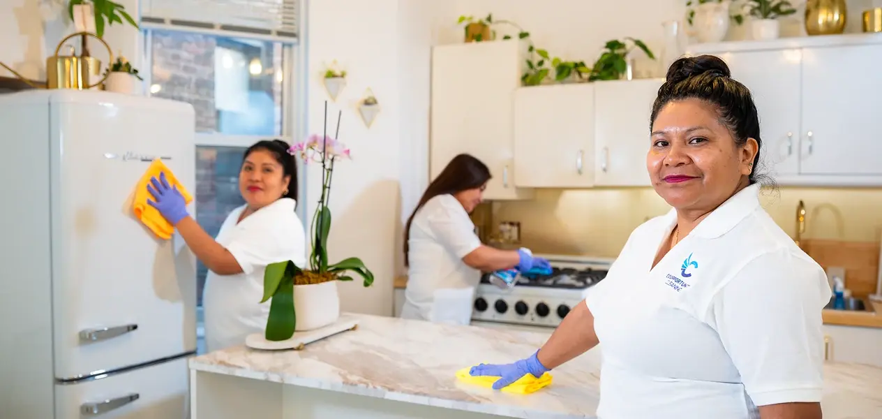 Professional female cleaners in white uniforms wiping down a kitchen refrigerator and counter, smiling and working together